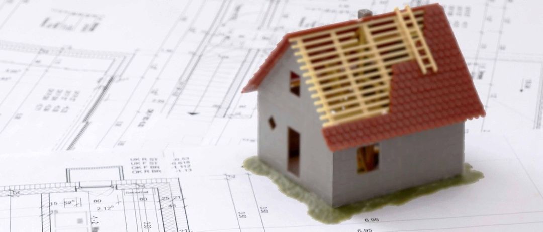 A house model rests atop architectural blueprints, accompanied by a yellow pencil, yellow measuring sticks, and a pair of pliers. The house model features a red roof, a ladder, and a small garden.