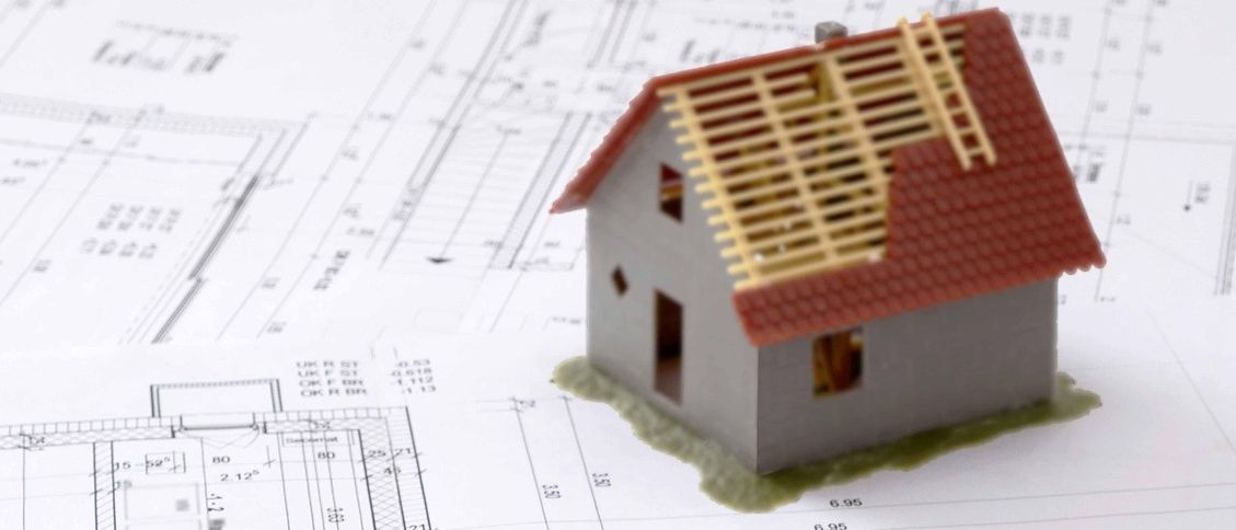 Ein Häusermodell ruht auf Architekturplänen, begleitet von einem gelben Bleistift, gelben Maßstäben und einer Zange. Das Häusermodell hat ein rotes Dach, eine Leiter und einen kleinen Garten.