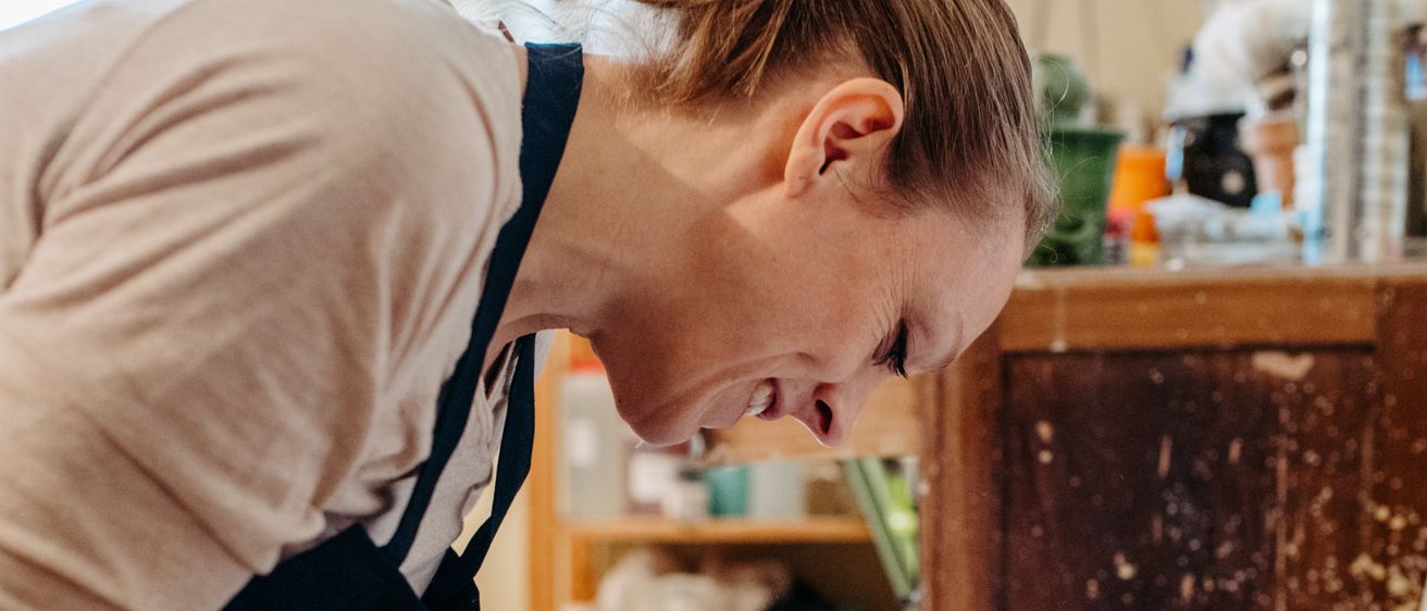 Eine Frau in einer Schürze arbeitet an einer Töpferscheibe und konzentriert sich auf ihre Aufgabe, mit tönerbedeckten Händen. Dahinter sind ein Holzschrank und Regale mit verschiedenen Gegenständen zu sehen.