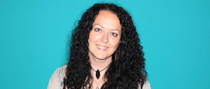 Eine Frau mit lockigem schwarzem Haar trägt ein schwarzes Oberteil und eine Halskette mit einem großen schwarzen Stein. Sie posiert vor einem blauen Hintergrund.