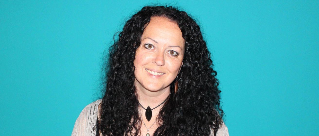 Eine Frau mit lockigem schwarzem Haar trägt ein schwarzes Oberteil und eine Halskette mit einem großen schwarzen Stein. Sie posiert vor einem blauen Hintergrund.