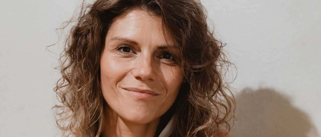 Eine Frau mit lockigem Haar lächelt und hält getrocknete Pflanzen. Sie trägt ein grünes ärmelloses Oberteil, ihr Arm ruht auf der Schulter. Der Hintergrund ist schlicht, möglicherweise drinnen.