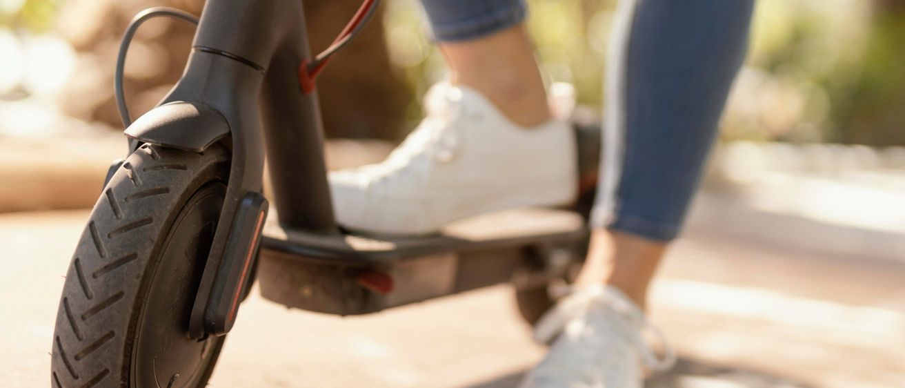 Eine Person in blauer Jeans und weißen Sneakern steht neben einem schwarzen Elektroroller, möglicherweise an einem sonnigen Tag.