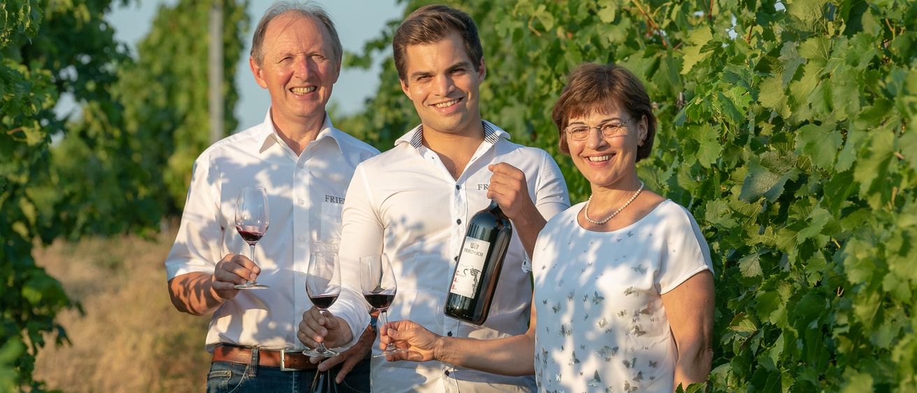 Drei Personen, zwei Männer und eine Frau, stehen in einem Weinberg und halten Weingläser und eine Flasche. Sie lächeln und posieren für ein Foto.