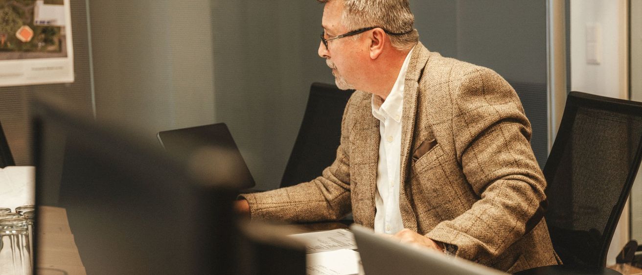 Ein älterer Mann mit Brille sitzt mit zwei Laptops an einem Schreibtisch und trägt einen braunen Mantel. Vor ihm liegen Papiere auf dem Schreibtisch.