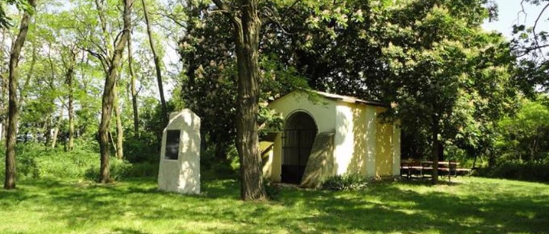 Eine kleine Kapelle mit einem gewölbten Eingang befindet sich in einem üppigen grünen Park. Sie ist von hohen Bäumen und einem Gedenkstein umgeben.
