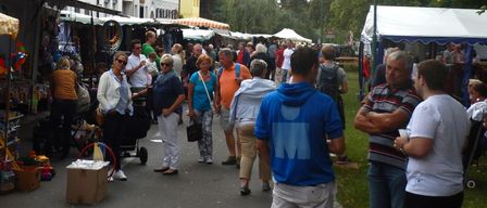 Eine Menschenmenge versammelt sich auf einem Freiluftmarkt. Ein Mann in einer blauen Kapuzenjacke schaut auf der linken Seite etwas an. Eine Frau mit Sonnenbrille schiebt einen Kinderwagen.