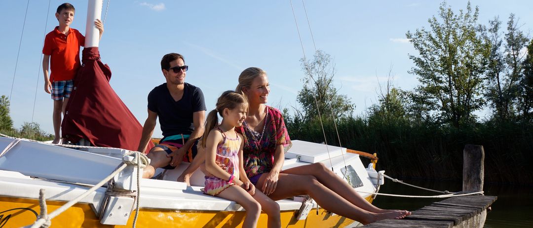 Eine vierköpfige Familie, bestehend aus einem Mann, einer Frau und zwei Kindern, sitzt lächelnd auf einem gelben Boot am Steg. Das Boot ist mit Seilen am Steg festgemacht.
