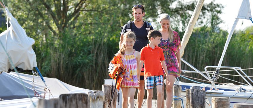 Eine Familie geht auf einem Holzsteg in der Nähe eines Bootes. Der Mann trägt einen Rucksack. Die Frau trägt ein Blumenkleid. Zwei Kinder, ein Mädchen und ein Junge, tragen Sommerkleidung. Bäume und Büsche sind im Hintergrund.