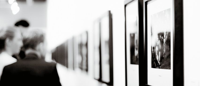 Eine Person betrachtet gerahmte Fotografien in einem schwach beleuchteten Raum. Die Bilder sind an einer weißen Wand angebracht.