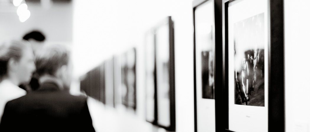 Eine Person betrachtet gerahmte Fotografien in einem schwach beleuchteten Raum. Die Bilder sind an einer weißen Wand angebracht.