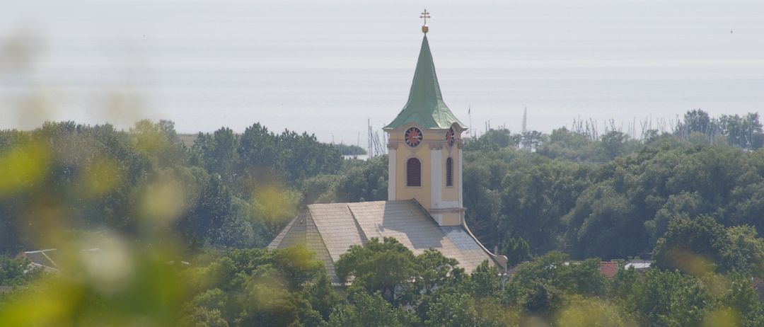 Eine Kirche mit einem Turm und einem Kreuz ist von Bäumen umgeben. Die Kirche hat eine Uhr auf dem Turm. Der Himmel ist trüb.