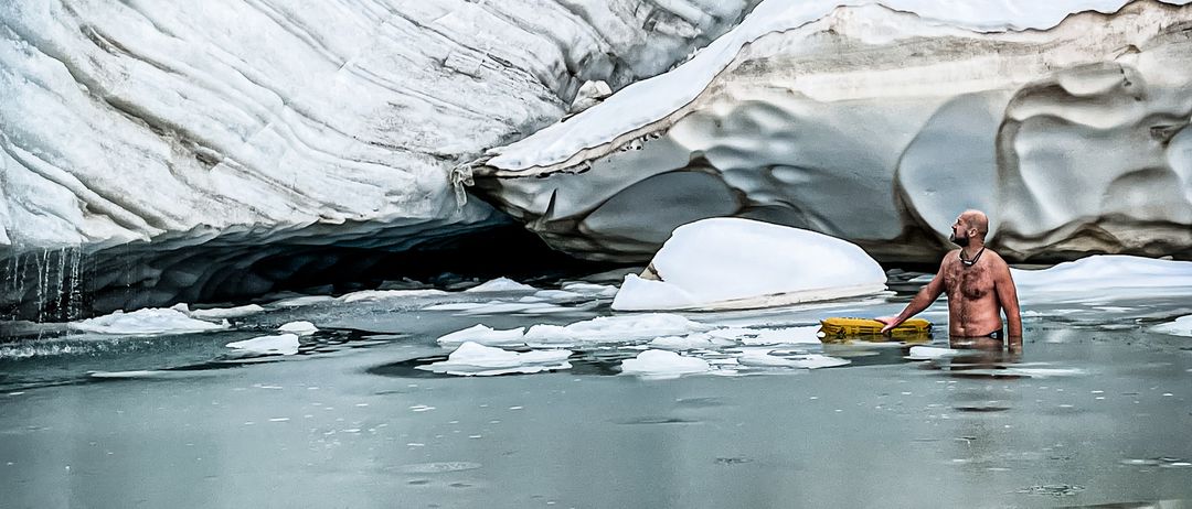 Ein Mann steht in eisigen Gewässern in der Nähe eines großen Gletschers und hält ein gelbes Objekt. Der Gletscher weist sichtbare Risse und Eisformationen auf.