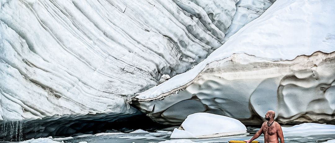 Ein Mann steht in eisigem Wasser in der Nähe eines Gletschers und hält ein gelbes Objekt, umgeben von Eis.