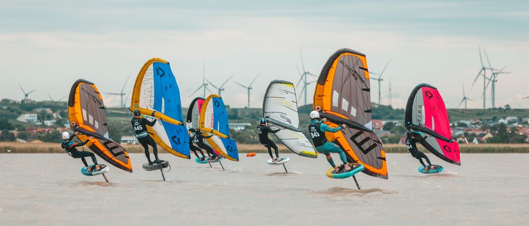 Eine Gruppe von Athleten auf Surfbrettern windsurft in einer Reihe auf ruhigem Wasser, mit bunten Segeln und Helmen, und Windturbinen in der Ferne.