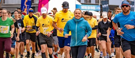 Eine Gruppe von Läufern nimmt an einem Marathon teil, trägt gelbe und blaue T-Shirts. Eine Frau in einer blauen Jacke führt die Gruppe an, gefolgt von einem Mann in einem schwarzen Hut. Im Hintergrund befinden sich weiße Zelte und Banner mit dem Wort 'LAUFEN' darauf.