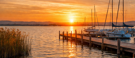 Ein friedlicher Sonnenuntergang über einem ruhigen See mit mehreren Segelbooten am hölzernen Steg verankert. Der Himmel ist mit warmen Farbtönen bemalt, und Reflexionen tanzen auf der Wasseroberfläche.