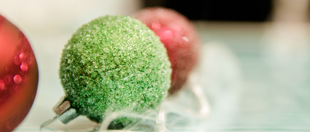 Ein Nahaufnahme eines grünen glitzernden Weihnachtskugels mit einem verschwommenen roten Ornament im Hintergrund auf einer gestreiften Oberfläche.