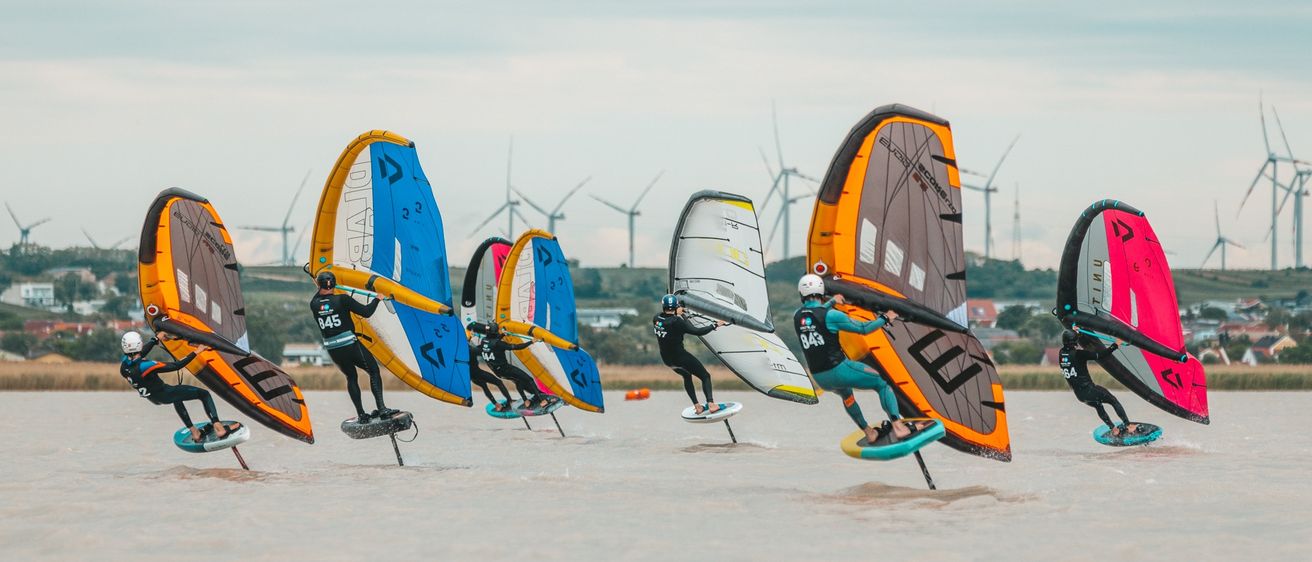 Eine Gruppe von Windsurfern gleitet mit Helmen und bunten Segeln über ruhiges Wasser. Im Hintergrund sind Windmühlen und ein klarer Himmel zu sehen.