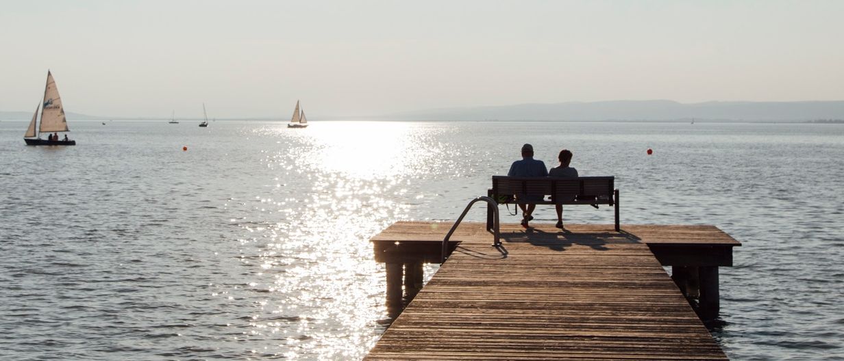 Zwei Personen sitzen auf einem Steg in Richtung Sonnenuntergang, mit Segelbooten in der Ferne. Das Wasser ist ruhig.