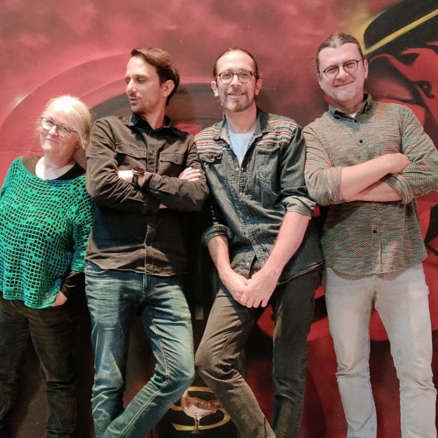 Vier Personen stehen nebeneinander vor einem roten und schwarzen Hintergrund mit einem Batman-Symbol.