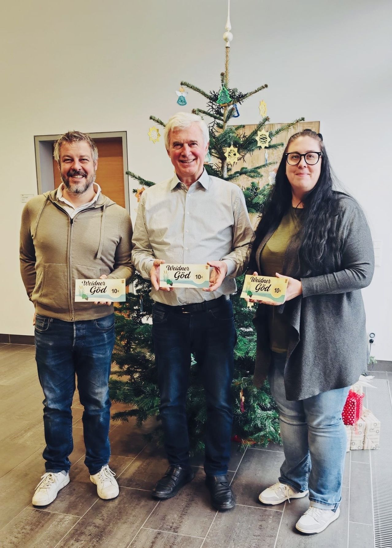 Drei Personen stehen vor einem Weihnachtsbaum. Der Mann links und die Frau rechts lächeln und halten eine Schachtel. Der Mann in der Mitte schaut zur Kamera. Dahinter ist eine weiße Wand.