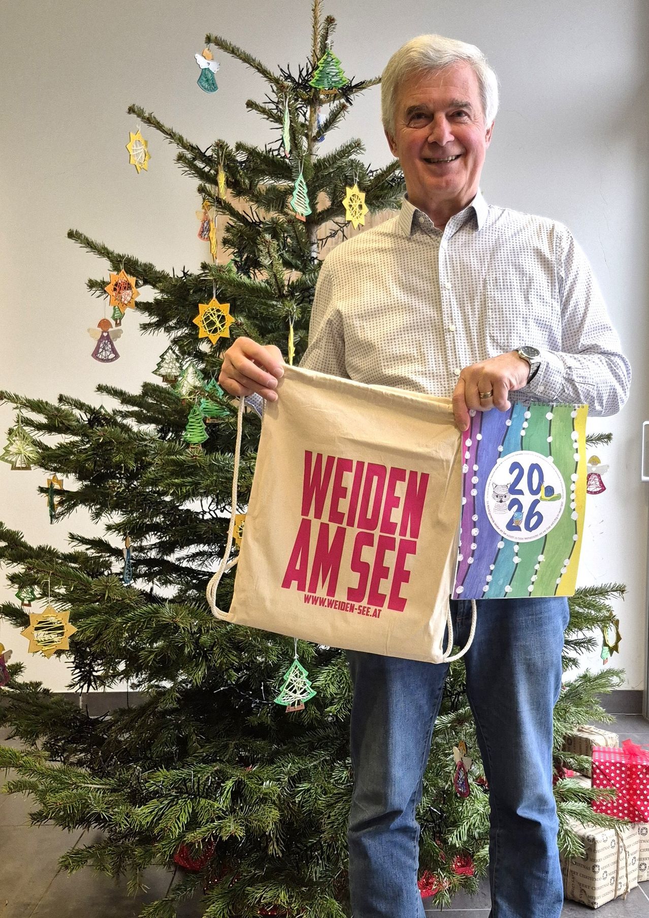 Ein Mann steht vor einem geschmückten Weihnachtsbaum und hält eine Tasche mit dem Text 'WEIDEN AM SEE' und ein buntes Buch. Er lächelt und trägt einen Ring.