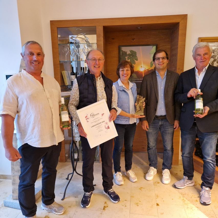 Fünf Personen stehen in einem Raum und lächeln für ein Foto. Der Mann auf der linken Seite hält eine Weinflasche, während der Mann in der Mitte ein Zertifikat in der Hand hat. Die Frau in der Mitte hält einen Pokal. Der Mann auf der rechten Seite hält eine Weinflasche. Hinter ihnen steht eine Glasvitrine.