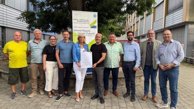 Eine Gruppe von Menschen steht vor einem Banner mit einem Baum und Gebäuden im Hintergrund. Sie lächeln und scheinen für ein Foto zu posieren.