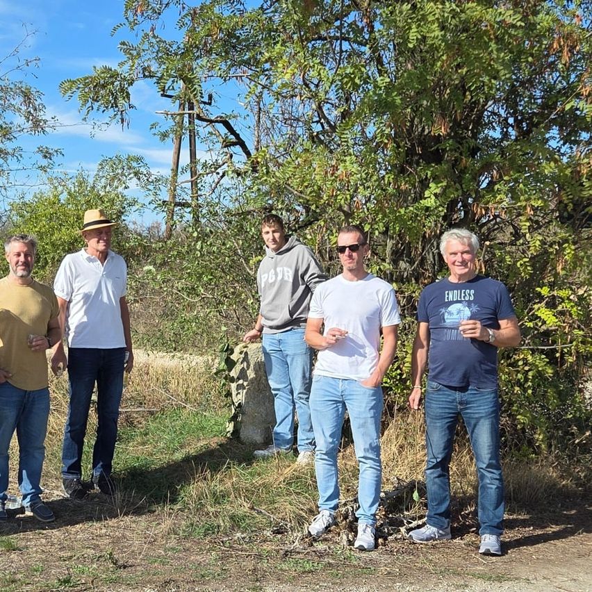 Fünf Männer stehen in einer Reihe unter einem Baum, alle in Freizeitkleidung. Ein Mann trägt einen Hut, und ein anderer hält ein Getränk. Der Hintergrund zeigt ein Grasfeld mit einer Steinstruktur.