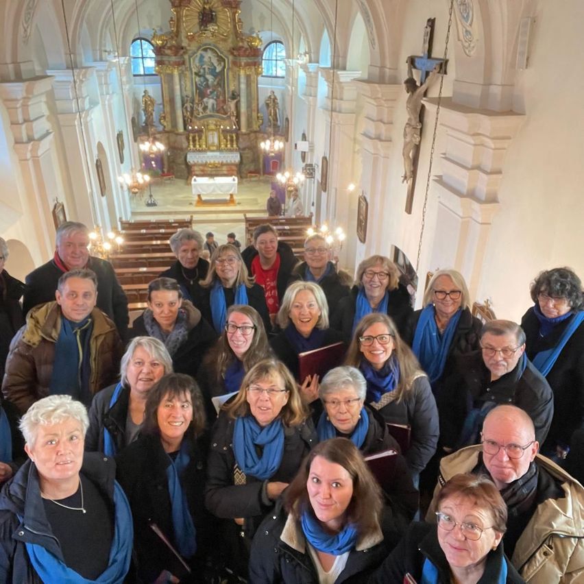 Eine Chorgruppe posiert für ein Foto in einer Kirche. Sie tragen blaue Schals und halten Bücher. Die Kirche hat einen goldenen Altar mit Statuen und einem Kreuz an der Wand.
