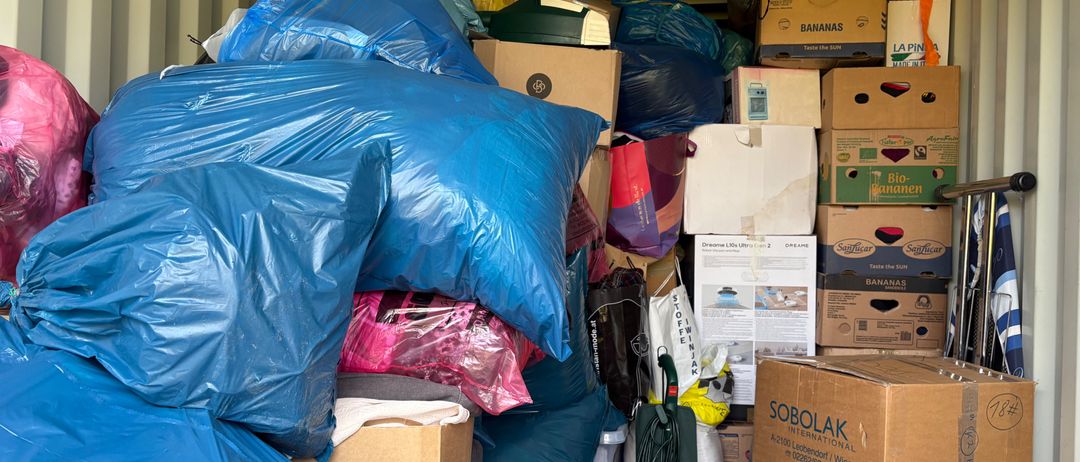 Ein Lagercontainer gefüllt mit verschiedenen Gegenständen, darunter blaue Plastiktüten, braune Kartons, ein lila Kissen und eine grüne Mülltonne. Einige Kartons haben Etiketten wie 'Bananen' und 'La Piña'.