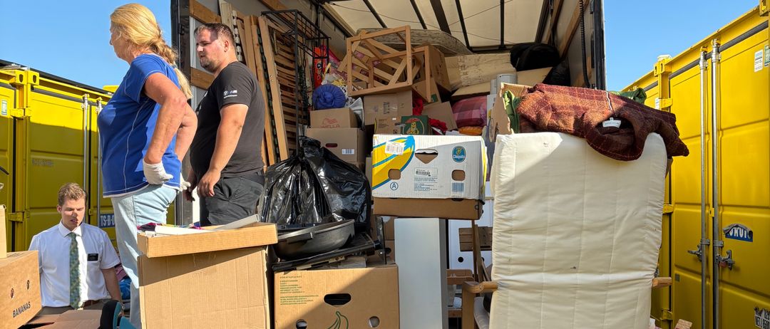 Zwei Personen laden Kisten in den Laderaum eines Lastwagens. Eine dritte Person hält die Tür offen. In der Nähe befinden sich weitere gelbe Lastwagen und Lagercontainer.