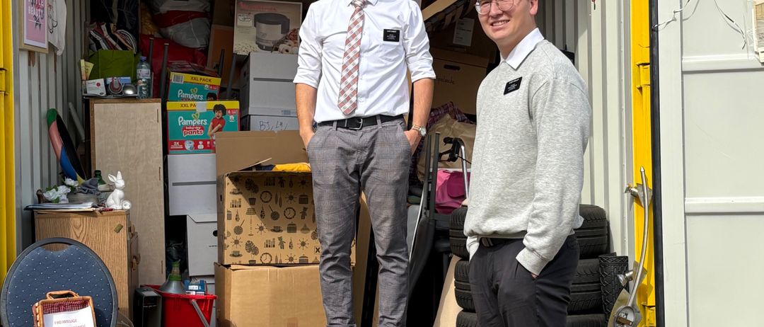 Zwei Männer stehen in einem mit verschiedenen Gegenständen gefüllten Container, darunter Kisten und Haushaltswaren. Ein Mann trägt Krawatte und Hut, der andere eine Brille. Links steht ein Stuhl mit einem Schild und einem Korb.