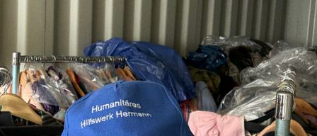 A man wearing a blue cap labeled 'Humanitäres Hilfswerk Hermann' stands in a storage container filled with clothes and plastic bags.