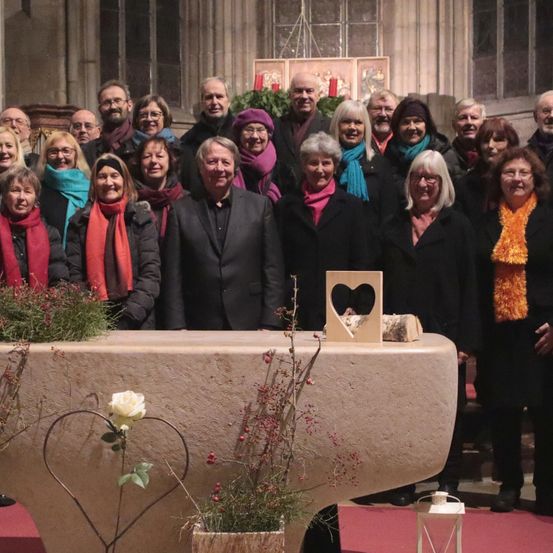 Eine Gruppe von Menschen posiert für ein Foto in einer Kirche, ein Mann in der Mitte trägt einen Anzug. Blumenarrangements und ein herzförmiger Holzrahmen sind im Vordergrund.