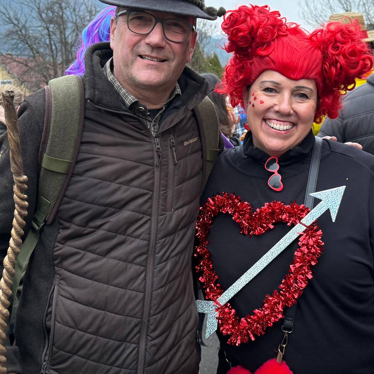 Ein lächelnder Mann und eine Frau mit roter Perücke posieren für ein Foto. Die Frau hat ein rotes herzförmiges Tinsel mit einem silbernen Pfeil. Der Mann hat einen Rucksack und einen Stock. Dahinter stehen mehrere Leute. Der Hintergrund ist unscharf.