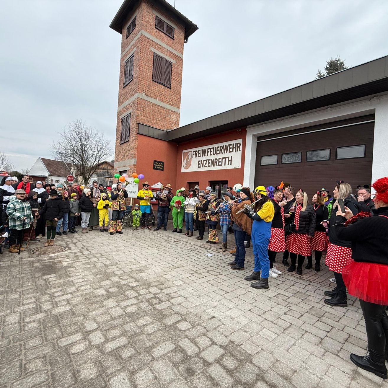 Eine Menschenmenge versammelt sich vor einer Feuerwehrstation, einige in Kostümen, mit einem Turm und Luftballons im Hintergrund.