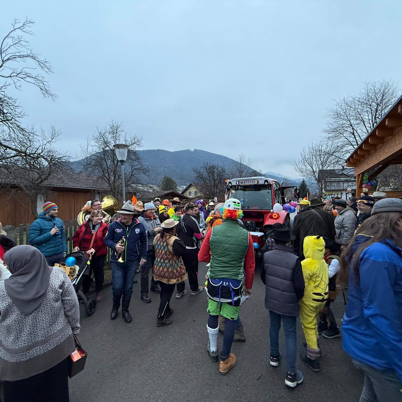 Eine Menschenmenge versammelt sich auf einem Dorfplatz, hinter ihnen parkt ein Traktor. Einige Leute tragen Kostüme, und einige tragen Musikinstrumente. Im Hintergrund sind Berge und Gebäude zu sehen.