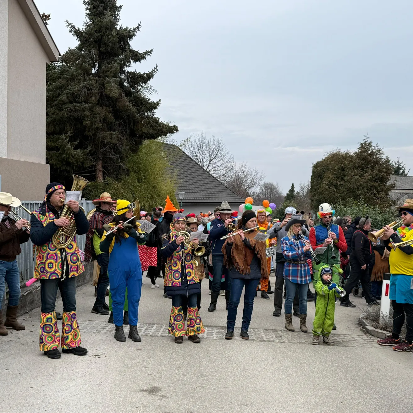 Eine Gruppe von Musikern in bunten Kostümen steht auf der Straße und spielt Blech- und Holzblasinstrumente, mit Zuschauern dahinter und Luftballons im Hintergrund.