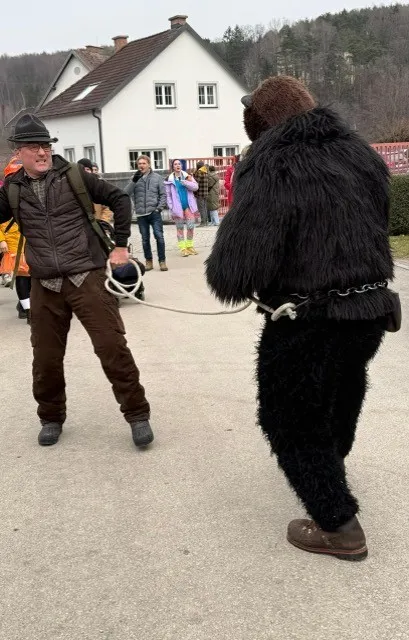 Ein Mann zieht ein Bärenkostüm, während andere hinter ihm zusehen. Im Hintergrund befindet sich ein Haus.
