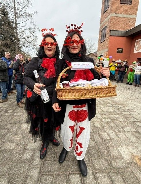 Zwei Personen in herzförmigen Kostümen halten einen Korb und eine Flasche und stehen im Freien. Dahinter Musiker und Zuschauer. Ein Backsteingebäude mit einem Schild ist im Hintergrund.