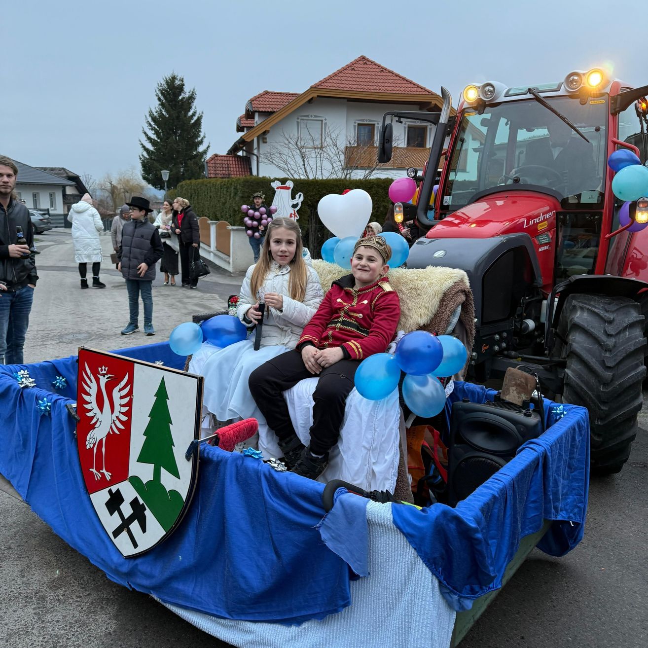 Zwei Kinder sitzen auf einem geschmückten Wagen, der von einem Traktor gezogen wird, mit einer Menge und Häusern im Hintergrund.