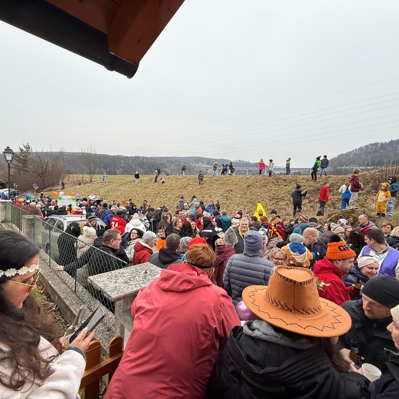 Eine große Menschenmenge in Winterkleidung versammelt sich auf einem Hügel, um eine Veranstaltung zu beobachten. Einige tragen Hüte, Brillen und halten Tassen. In der Ferne sind Menschen am Boden, eine Brücke und eine Bergkette zu sehen.