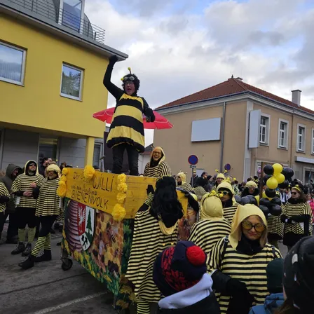 Eine Gruppe von Menschen in Bienenkostümen zieht in einem Umzug, wobei eine Person in einem Bienenkostüm auf einem float steht. Der float hat ein gelbes und schwarzes Design. Dahinter sieht eine Menge zu. Im Hintergrund befinden sich Gebäude und Luftballons.