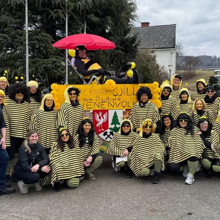 Eine Gruppe von Menschen in gelben und schwarzen Kostümen, die lächelnd für ein Foto posieren. Dahinter ein gelber Banner mit einem roten Regenschirm und Luftballons. Ein Haus mit Schornstein und Bäumen im Hintergrund.