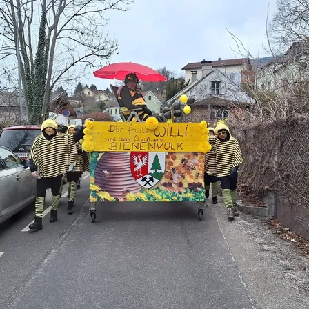 Eine Gruppe von Menschen in Bienenkostümen zieht auf einem Wagen mit einem Mann, der unter einem roten Regenschirm sitzt. Der Wagen hat ein gelbes Schild mit den Worten Der Faule Willi und sein Bienes BieneNvolk.