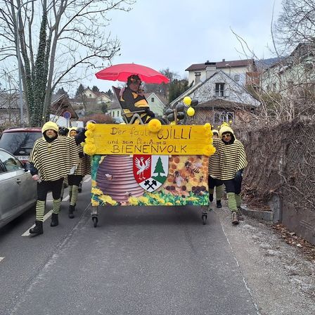 Eine Gruppe von Menschen in Bienenkostümen zieht auf einem Wagen mit einem Mann, der unter einem roten Regenschirm sitzt. Der Wagen hat ein gelbes Schild mit den Worten Der Faule Willi und sein Bienes BieneNvolk.