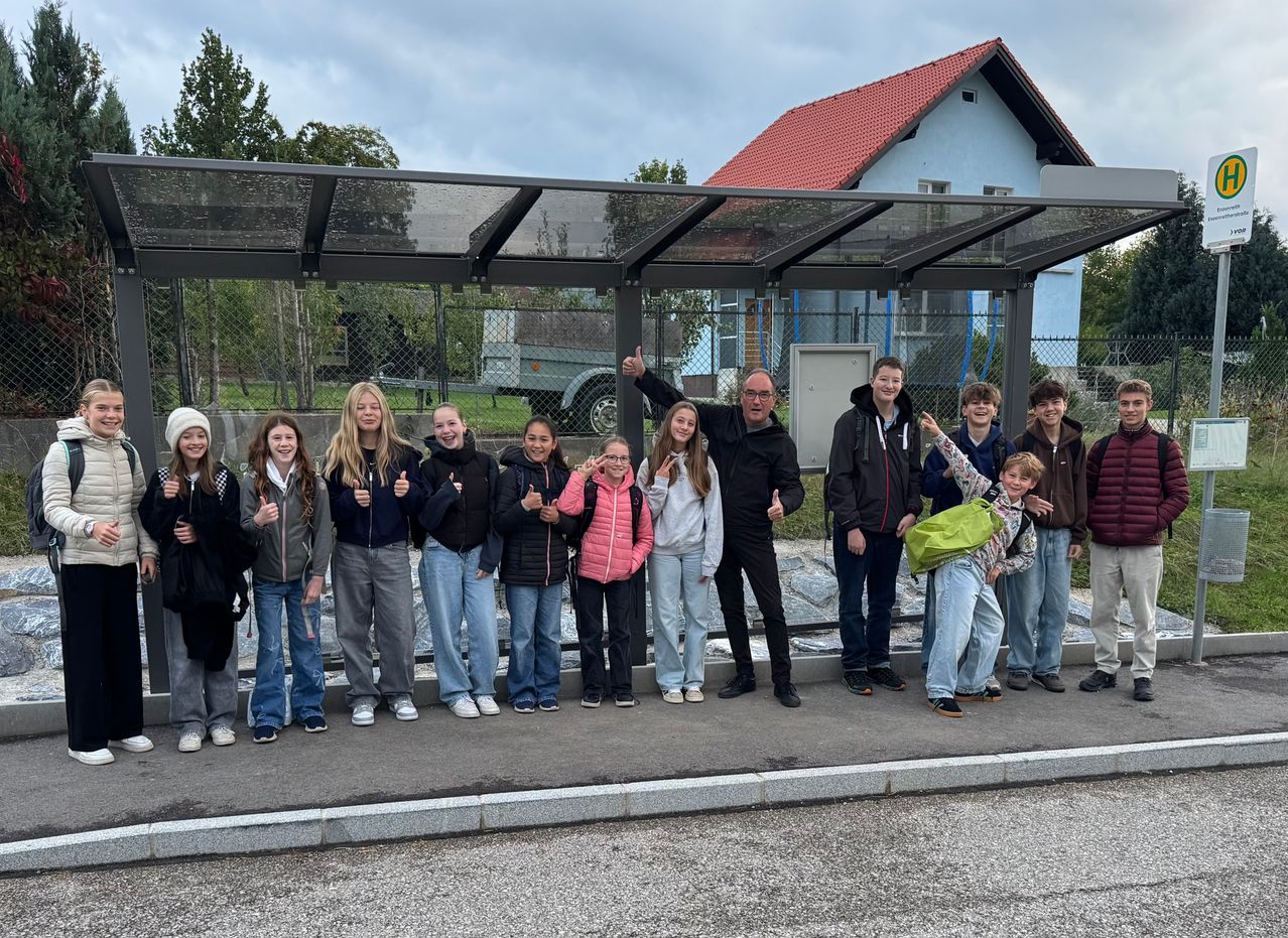Eine Gruppe von Schülern und zwei Männern posiert für ein Foto vor einer Bushaltestelle, mit einem Haus und Grünflächen im Hintergrund.