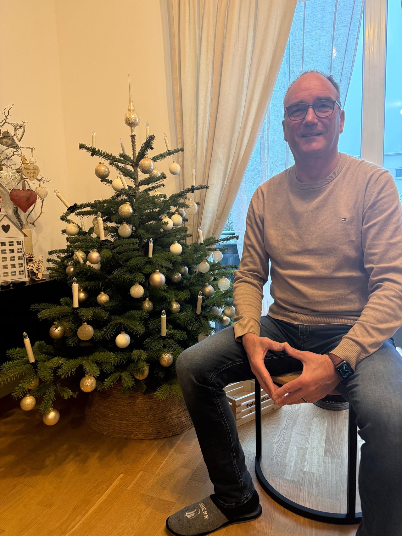 Ein Mann sitzt auf einem Hocker vor einem geschmückten Weihnachtsbaum. Er trägt eine Brille und einen beigen Pullover, seine Hände sind ineinander verschränkt. Der Baum ist mit Ornamenten und Kerzen geschmückt. Hinter ihm ist ein Fenster mit Vorhängen zu sehen.